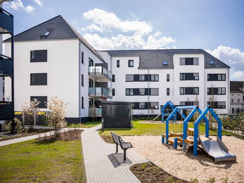 Rückansicht des Gebäudes, vorne rechts der Spielplatz, links daneben ein Weg mit Bank und Rasen.