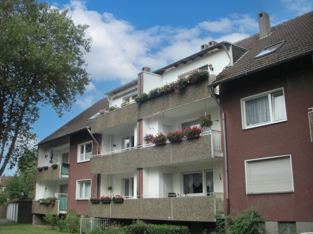 Ein zweistöckiges Mehrfamilienhaus mit rot-brauner Fassade und Balkonen mit grauer Betonfassade. Blauer Himmel mit Wolken, links im Anschnitt ein Baum.
