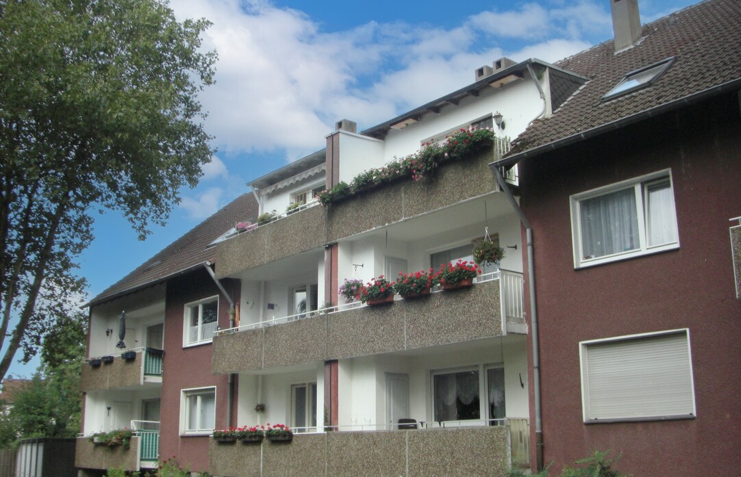 Ein zweistöckiges Mehrfamilienhaus mit rot-brauner Fassade und Balkonen mit grauer Betonfassade. Blauer Himmel mit Wolken, links im Anschnitt ein Baum.