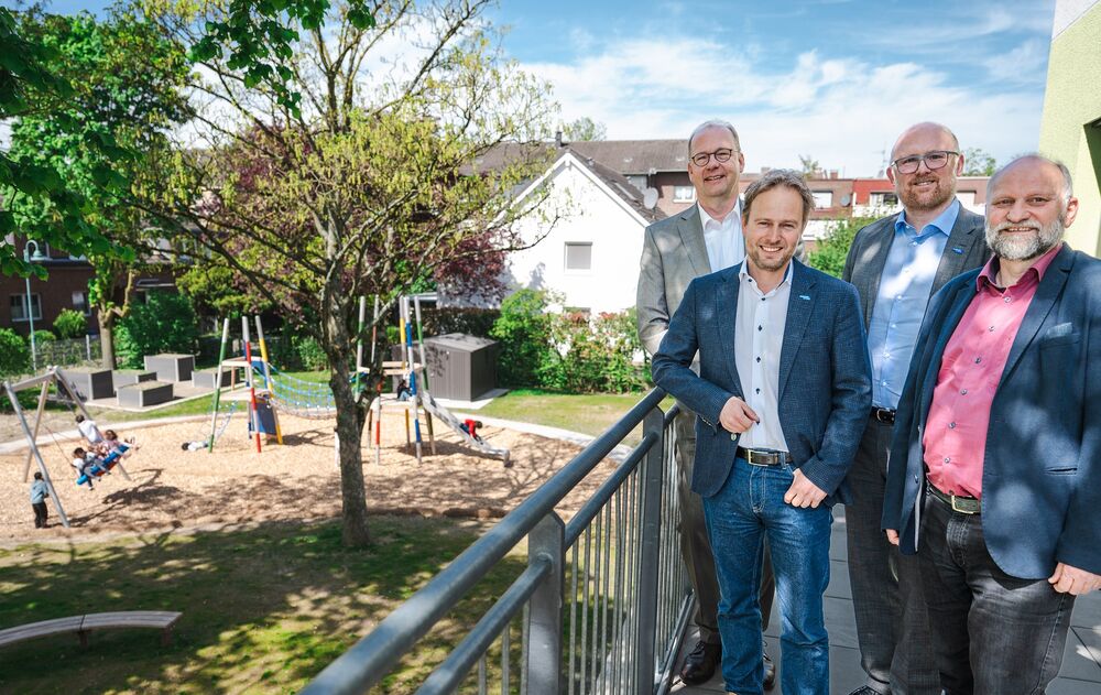 Vier Männer stehen beieinander an einem Geländer. Links neben Ihnen spielen Kinder auf dem Spielplatz.
