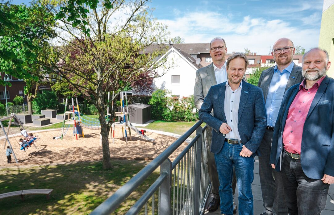 Vier Männer stehen beieinander an einem Geländer. Links neben Ihnen spielen Kinder auf dem Spielplatz.