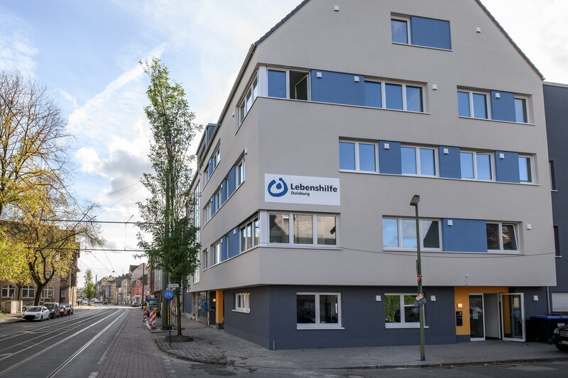 Ansicht des Neubaus von der Straße aus, das Haus steht an einer Ecke. Breite Fensterfronten, das Logo der Lebenshilfe auf der Fassade.