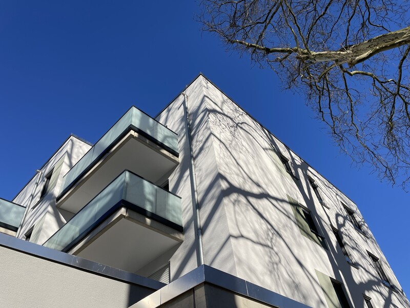 Ein Wohngebäude mit weißer Fassade vor blauem Himmel, ein Baum rechts wirft einen Schatten auf das Haus. Auskragende Milchglasbalkone.