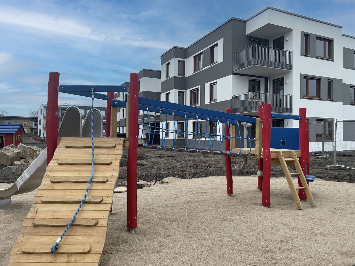 Ein Spielplatz mit Rutsche und Kletternetz vor einem modernen dreistöckigen Wohngebäude. Der Außenbereich ist unbepflanzt.